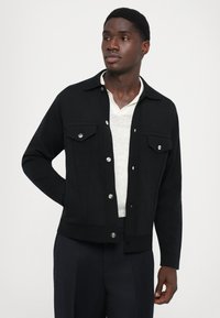 Černá bunda z hladké látky, s dvěma náprsními kapsami, stříbrnými knoflíky, límečkem a zkráceným střihem. Nositá přes světlý top.