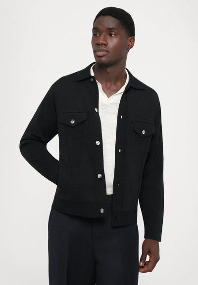 Sort button-up jakke i en bomuldsblanding, med to brystlommer, krave og kort design. Båret over en lys skjorte.