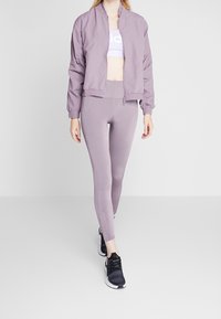 Ljuslila bomberjacka, figurnära ljuslila leggings och svarta träningsskor. Outfiten har en sportig design med dragkedja.