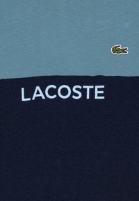 Bavlněné tričko s dvoubarevným designem v světle modré a námořnické. Uprostřed bílý nápis "LACOSTE" s malým zeleným krokodýlem v levém horním rohu.