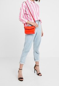 Person iført en lyserød og hvid stribet skjorte, lyse opsmøgede jeans, sorte højhælede sandaler og bærende en rød crossbody taske.