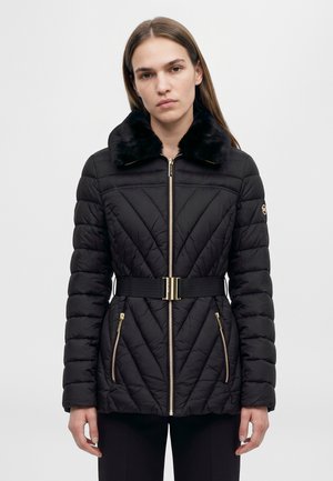 Mannequin portant un manteau noir matelassé avec un col en peluche, une fermeture éclair et des poches dorées, ainsi qu’une ceinture, sur un fond clair uni.
