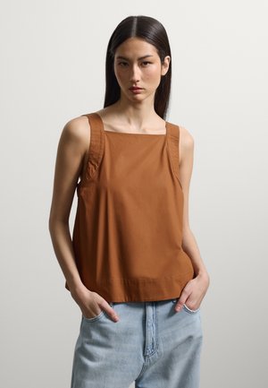 Femme aux longs cheveux foncés portant un haut marron sans manches et un jean bleu clair, mains dans les poches, fond neutre.