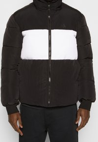 Chaqueta acolchada en blanco y negro con cremallera frontal y puños acanalados, llevada por una persona que muestra el torso y las manos.