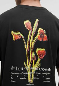 Tricou negru cu un grafic floral vibrat, pixelat, în nuanțe de roșu și galben, având text dedesubt în alb, creând un contrast vizual îndrăzneț.
