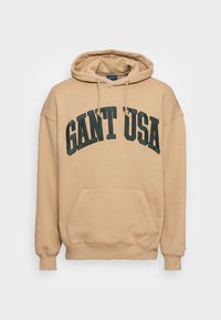 Beżowa bluza z kapturem, z przednią kieszenią i sznurkami, z dużym, ciemnozielonym napisem "GANT USA" na klatce piersiowej.