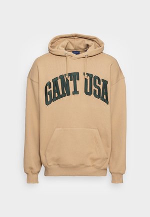 Beige mikina s kapucí, přední kapsou a šňůrkami, s velkým tmavě zeleným textem "GANT USA" přes hruď.