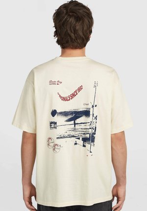 Man wearing cream t-shirt with graphic of a surfer and ocean, red text "Originals Since 1952" and "Santa Cruz California" on back.