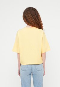 Κίτρινο πουλόβερ με κοντά μανίκια, χαλαρή εφαρμογή και στρογγυλή λαιμόκοψη, σε συνδυασμό με ανοιχτόχρωμο τζιν σε στενή γραμμή, προβολή από πίσω.