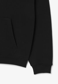 Detalle de la manga de sudadera negra y bolsillo tipo canguro sobre un fondo blanco liso, mostrando un suave puño acanalado y una costura limpia.