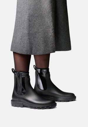 Personne portant des bottes de pluie noires chevillères avec des fermetures éclair latérales et des tirettes, des collants noirs transparents et une jupe grise longueur genoux, debout sur un fond clair.