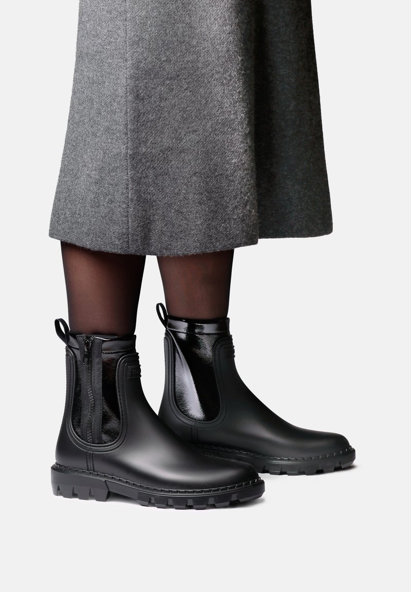 Personne portant des bottes de pluie noires chevillères avec des fermetures éclair latérales et des tirettes, des collants noirs transparents et une jupe grise longueur genoux, debout sur un fond clair.