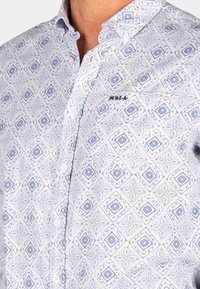 Blauw en wit gestreept shirt met een geometrisch ontwerp, knoopsluiting, en geborduurd logo op de borst. Glad stoftextuur.