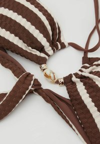 Maillot de bain texturé à rayures marron et crème avec détail de perles dorées et en marbre sur le devant, noué sur un fond blanc.