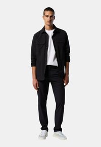 Camicia nera con bottoni e due tasche sul petto, indossata sopra una T-shirt bianca, abbinata a pantaloni neri e sneaker chiare.