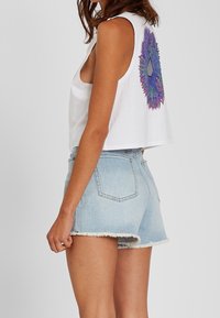 Top corto blanco con un gráfico circular en morado y azul en la parte trasera, combinado con pantalones cortos de mezclilla azul claro con bordes deshilachados.