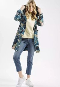 Lekka, kapturowa kurtka przeciwdeszczowa w niebiesko-zielony tropikalny wzór liści, w zestawieniu z beżowym swetrem i niebieskimi jeansami ściętymi na długość 7/8. Białe sneakersy.