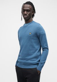 Modro dolg rokav pulover z okroglim izrezom, s majhnim zlatim emblemom ptice na prsih. Mehka tekstura, prilegajoč se kroj in rebrast spodnji rob.