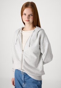 Svijetlosiva zip-up dukserica s kapuljačom na vezanje, slobodnog kroja, meke teksture i rebrastim manžetama. Nosi se preko krem topa i plavih traperica.