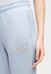 Ανοιχτό μπλε φούτερ παντελόνι με πλαϊνή τσέπη και χρυσό λογότυπο EA7 Emporio Armani από μικρά καρφάκια στον μηρό, χέρι που ακουμπάει δίπλα στο παντελόνι.