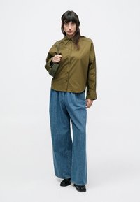 Πουκάμισο σε ελιάς με κουμπιά και γιακά, σε συνδυασμό με φαρδιά μπλε τζιν παντελόνια. Το μοντέλο κρατά μια μικρή πράσινη τσάντα και φοράει μαύρα παπούτσια.