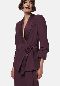 Donna che indossa un blazer in satin di un profondo colore prugna con un fiocco in vita e pantaloni abbinati, in posa su uno sfondo bianco.