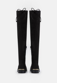 Bottes noires en daim au-dessus du genou avec des bouts arrondis, un laçage à l'arrière et des fermetures éclair latérales. Semelles plates avec un petit accent métallique au talon.