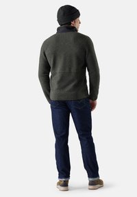 Mörkgrön ulltröja med svart krage, i kombination med blå jeans och tan färgade sneakers. Hatten är mörk och åtsittande. Synliga sömmar och texturdetaljer.