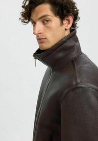 Brun läderjacka med hög krage, utrustad med en främre dragkedja och slät yta. Strukturerad design med subtila sömmar.