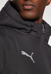 Sort polstret jakke med høj krave, der har et sølv Puma-logo på brystet og detaljer med frontlynlås. Glat overfladetextur.