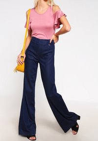 Pink off-the-shoulder top with ruffled sleeves, high-waisted navy pants, and black heels, carrying a yellow crossbody bag with a tassel.
