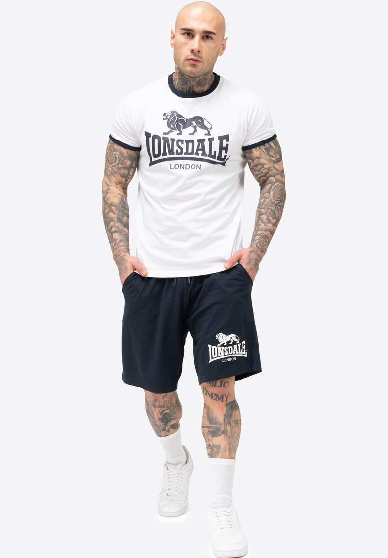 Valkoinen puuvillapaita mustilla saumoilla ja Lonsdale-logolla; yhdistettynä tummansinisiin shortseihin, joissa on logo. Mallilla on päällään valkoiset lenkkitossut ja valkoiset sukat.