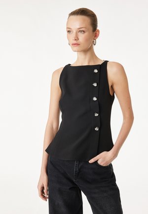 Kobieta ubrana w czarny top bez rękawów z kwadratowym dekoltem i rzędem pięciu guzików w kształcie serc z przodu, w połączeniu z czarnymi spodniami.