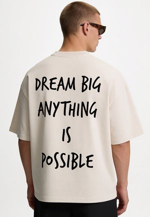Man draagt een zonnebril en een beige shirt met zwarte tekst op de achterkant waarop "DREAM BIG ANYTHING IS POSSIBLE" staat, tegen een witte achtergrond.