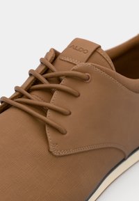 Zapatos marrones con cordones, compuestos de un material sintético texturizado con un acabado suave, que presentan cordones en un tono similar y un sutil logo en la lengüeta.