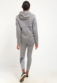 Žena odchází oblečená v šedé mikině s kapucí a šedých legínách Nike, na sobě má černé tenisky, na čistě bílém pozadí.