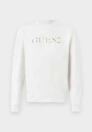 Balta garo piedurkņu kreklā ar apstrādātu logo "GUESS EST. 1981" uz krūtīm, attēlots uz vienkārša baltā fona.