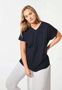 Marineblaue V-Ausschnitt-T-Shirt mit kurzen, breiten Ärmeln und lockerer Passform, kombiniert mit weißen Hosen. Glatter Stoff mit minimaler Gestaltung.