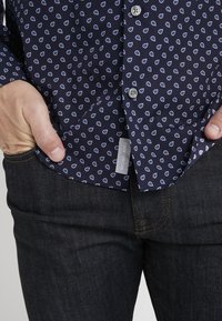 Camicia blu navy con un piccolo motivo a goccia bianca, presenta un'etichetta con il nome del marchio, abbinata a jeans in denim scuro.