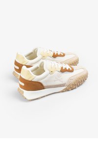 Sneaker aus einer Kombination von beigem und braunem Wildleder und Leder, mit einer strukturierten Sohle und einem Totenkopf-Logo an der Seite.