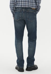 Žmogus, vilkintis mėlynus džinsus su užpakalinėmis kišenėmis ir juodomis batais, stovi prieš šviesų vientisą foną.