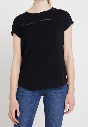 Kvinde iført en sort kortærmet top med en dekorativ vandret trim nær brystet og blå jeans.
