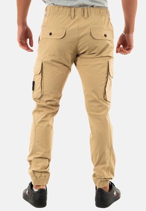 Homme portant un pantalon cargo beige avec des poignets élastiques et des baskets noires, debout de dos sur un fond blanc uni.