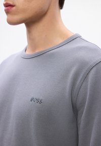 Siva strukturirana majica s dugim rukavima i okruglim izrezom, sa izvezenim logotipom "BOSS" na prsima. Materijal izgleda udobno i neformalno.
