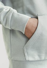 Felpa grigia chiaro con polsini a coste e tasca frontale, dal tocco morbido. Il design è semplice e funzionale.