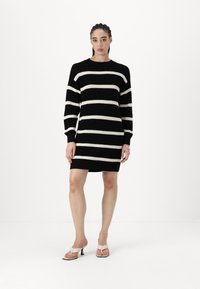 Vestido de malha listado em preto e branco com mangas longas e decote redondo. A bainha termina acima do joelho. Usado com sandálias de salto brancas.