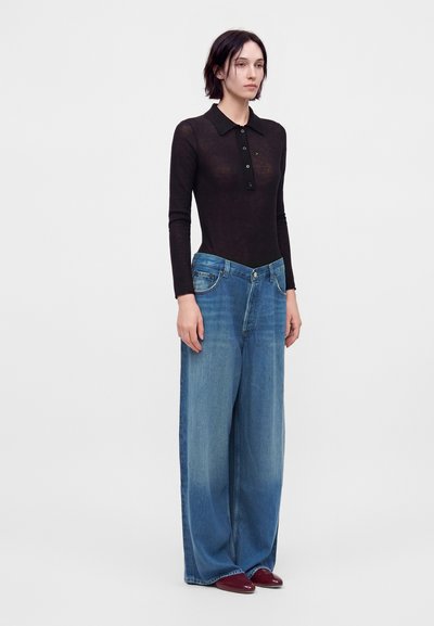 Femme portant un polo noir transparent à manches longues, un jean bleu large et des chaussures rouges, debout devant un fond blanc uni.
