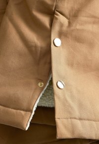 Brun stofjakke med en blød foring, glatte metalknapper og en tekstureret overflade med et subtilt vævemønster.