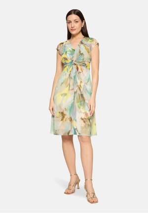 Frau trägt ein knielanges, kurzärmeliges Kleid mit floralem Muster und gedrehtem Vorderteil, kombiniert mit goldenen, hochhackigen Riemchensandalen.