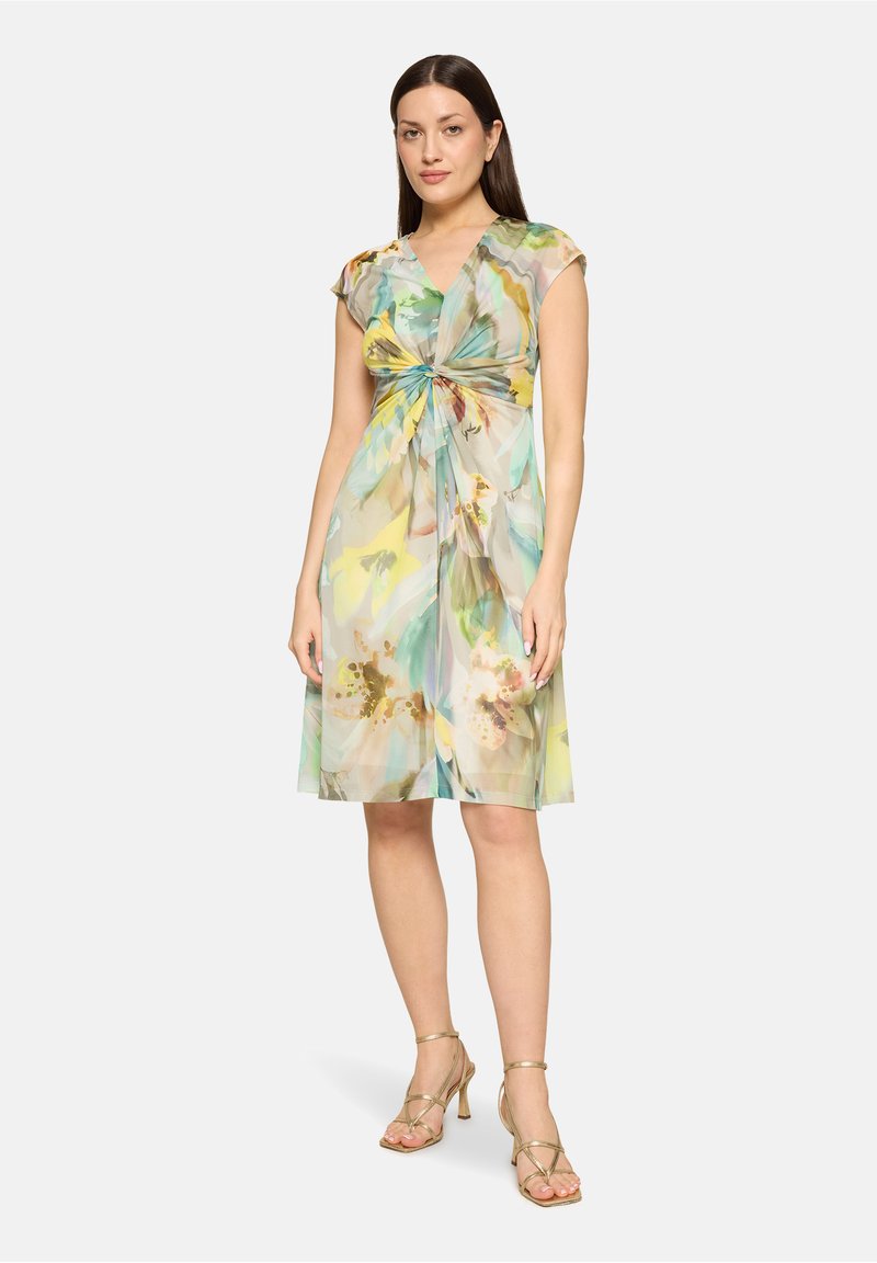 Femme portant une robe fleurie courte aux manches courtes avec un détail twisté à l'avant, associée à des sandales à talons hauts dorées avec des brides.